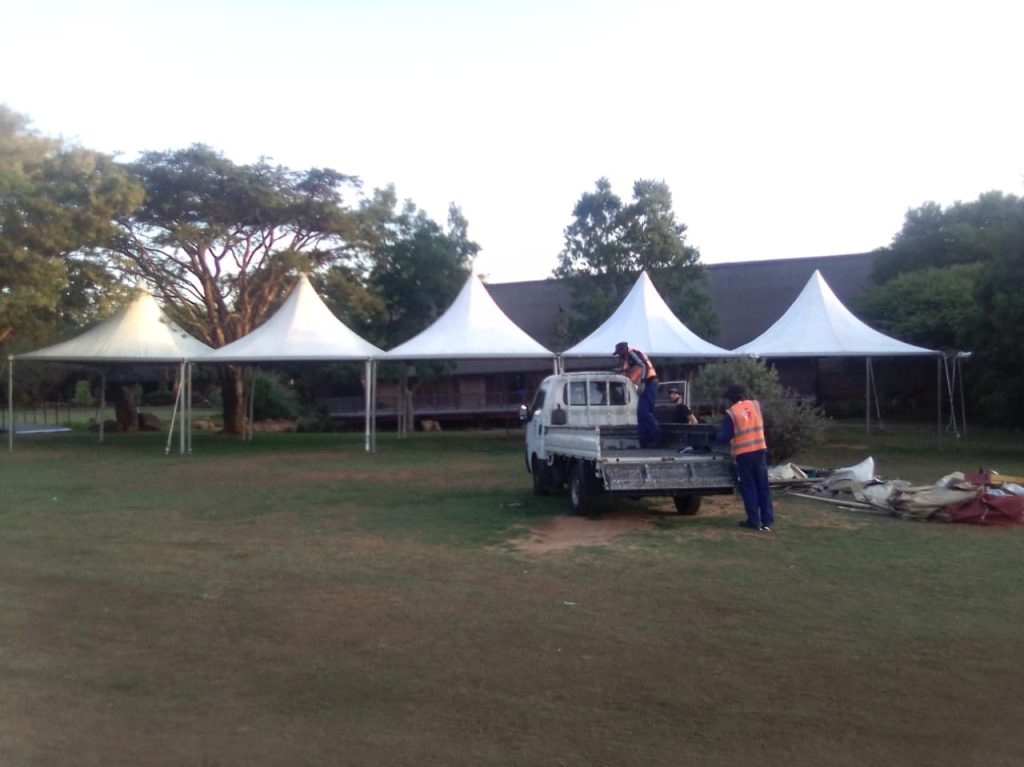 Events Guys Workers in orange safety vests set up white tents on a grassy field, using a pickup truck for assistance—ideal for anyone searching for "tent hire near me.